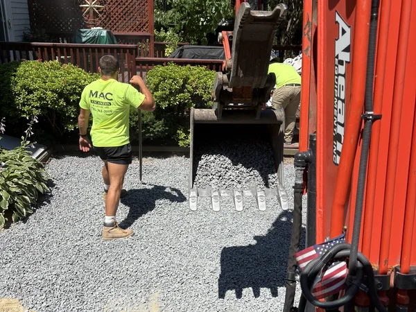 Crew spreading crushed gravel base with a mini excavator during paver hardscape installation in East Bridgewater, MA