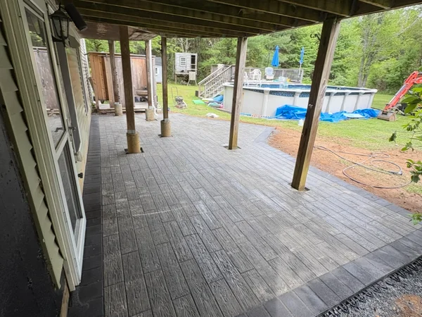 Wood-grain stamped paver patio with dark border installed under a deck beside a pool in Kingston, MA